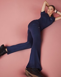 Woman in navy blue scrubs lounging with pink background.